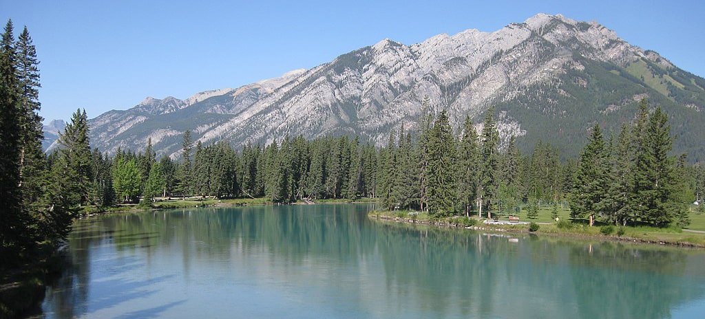 A serene mountain lake with clear turquoise waters surrounded by lush green trees and rugged peaks under a blue sky, Westhills, AB.