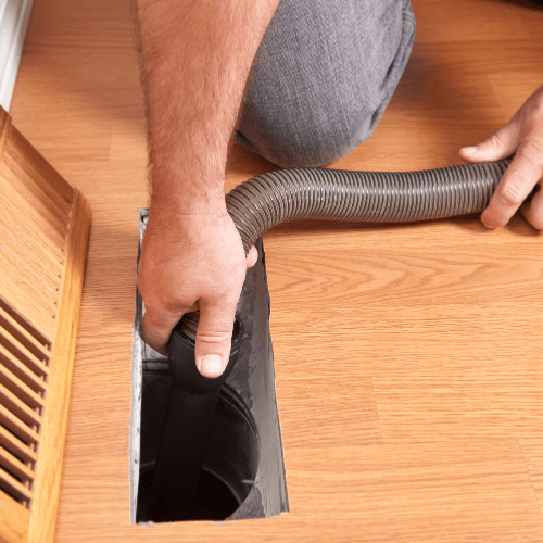 Person cleaning a vent on the floor with a flexible vacuum hose.