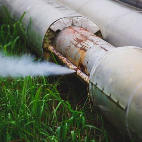 Rusty industrial pipes with steam leaking through a joint, surrounded by grass in Calgary.